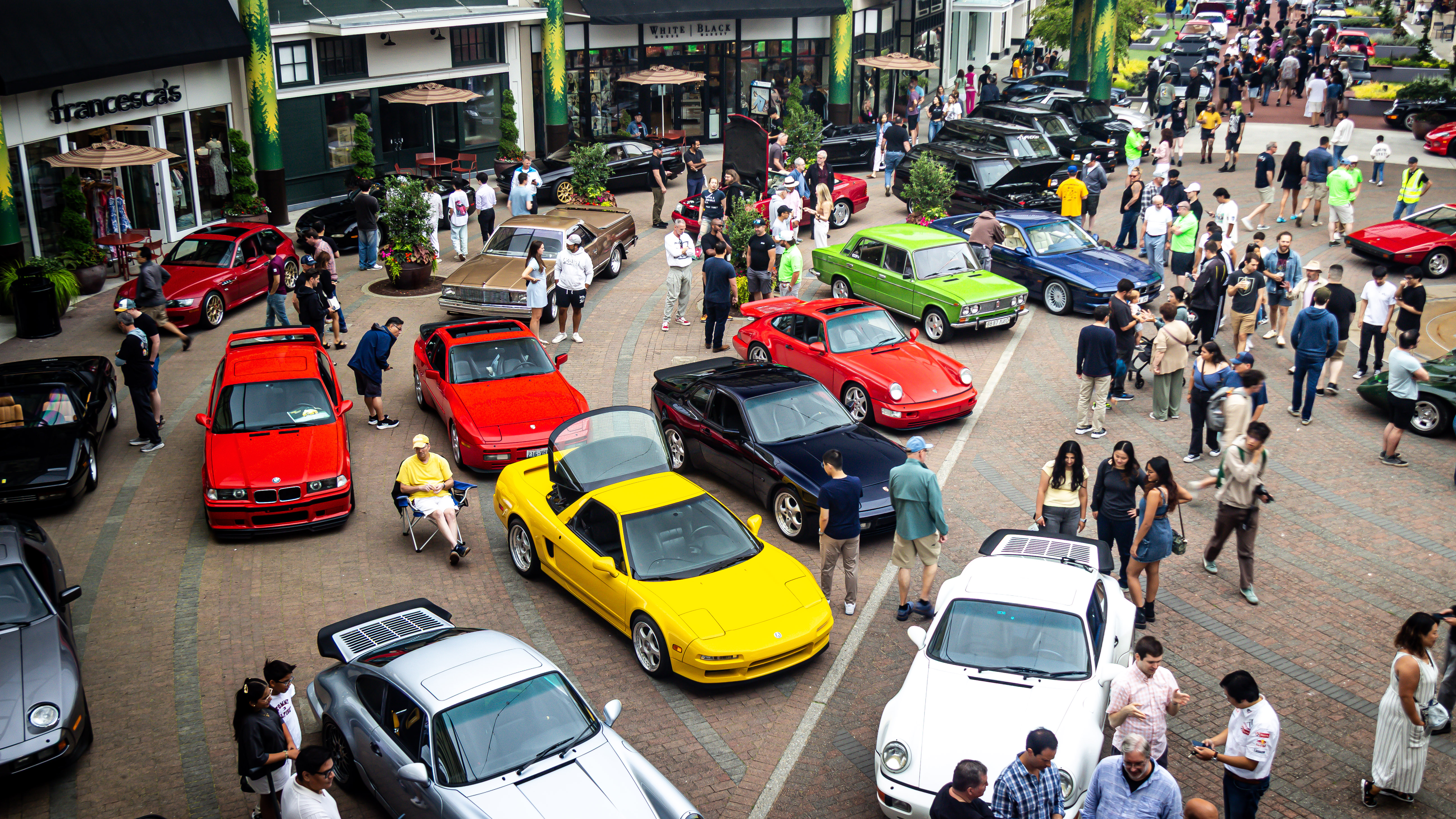 Aerial view of a large outdoor car show with colorful cars including yellow NSX, red BMW E36, and Porsches surrounded by crowds of people
